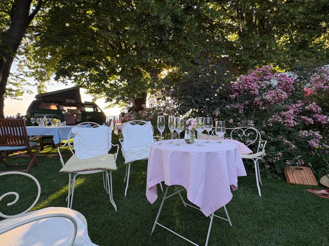 Gartenfest: Gartentisch mit Tischdecke und Sektgläsern, Stühlen  mit Kissen mit einem Bullybus als Cafebar im Hintergrund im Abendlich vor einem Rosenstrauch