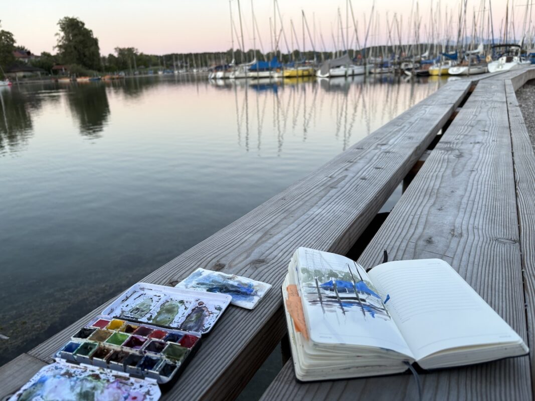 am Chiemsee. Ausblick auf das Wasser mit Aquarellkasten udn aufgeschlagenem Skizzenbuch mit gemalten booten im vordergrund
